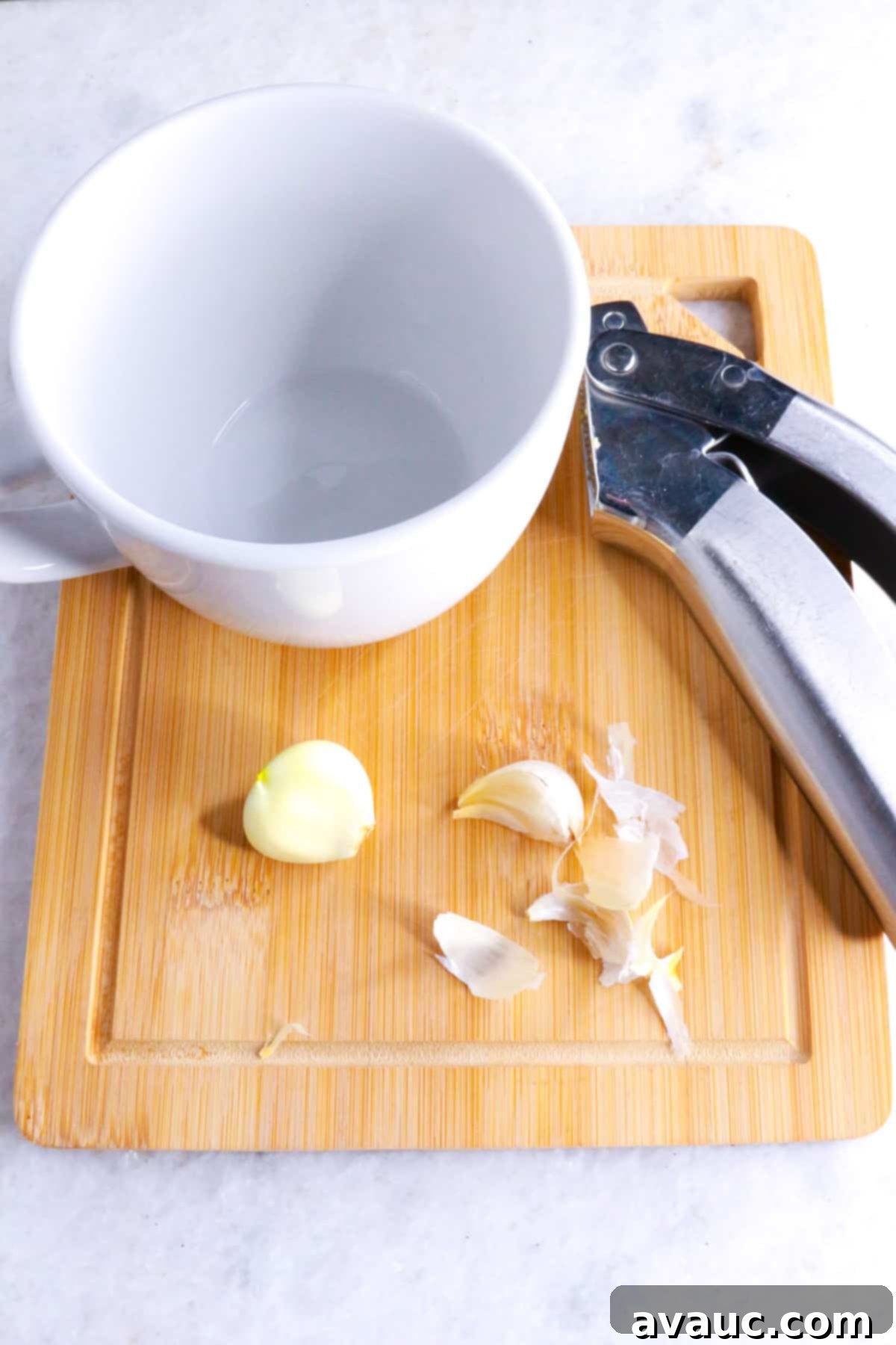 Homemade Garlic Tea Elixir 4 Close-up of a white mug, a metal garlic press, and several unpeeled garlic cloves resting on a light wooden cutting board, preparing for tea.
