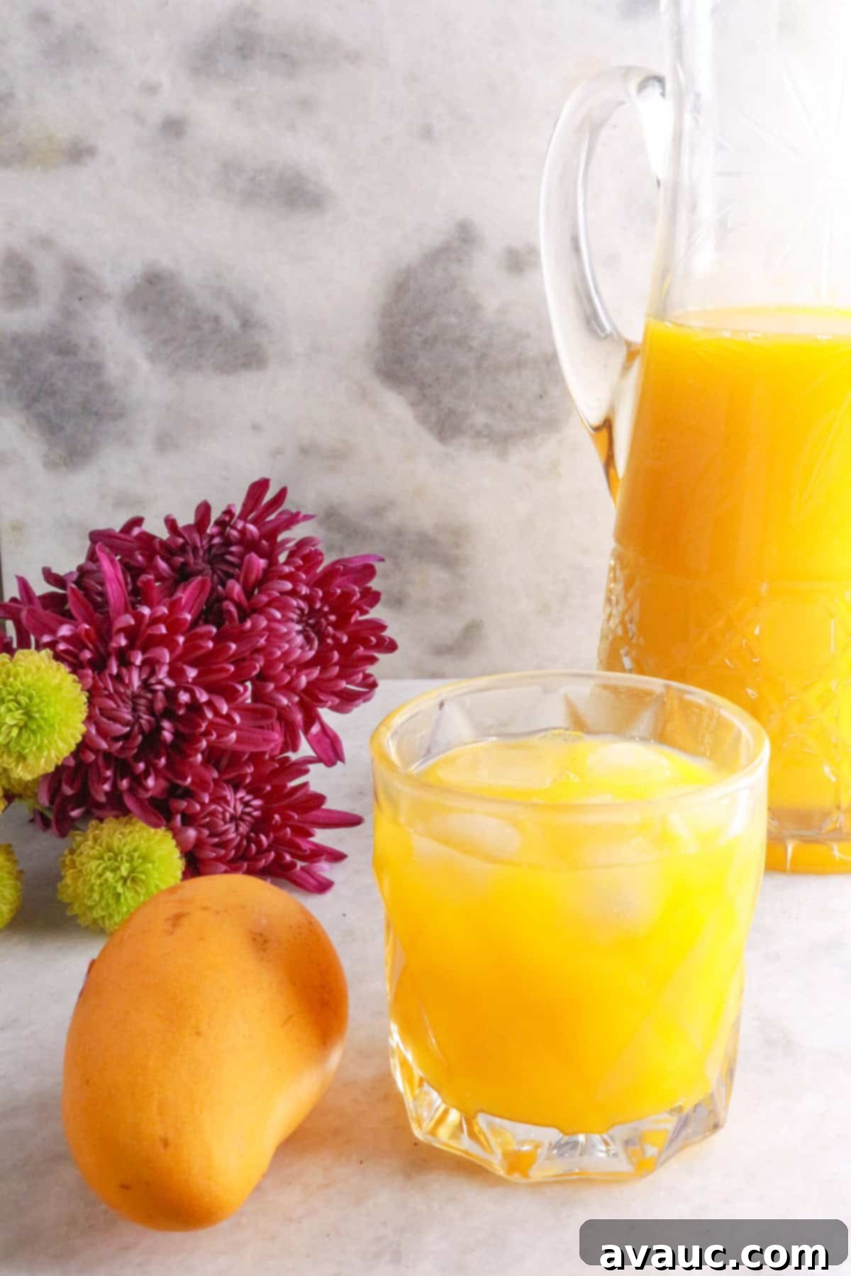 Simple Refreshing Mango Agua Fresca 2 Clear glass full of mango agua fresca with a yellow mango beside it and flowers and a glass pitcher of mango water behind it.