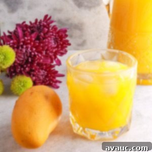 Simple Refreshing Mango Agua Fresca 10 Glass full of mango agua fresca with a yellow mango beside it and flowers and a glass pitcher of mango water behind it.