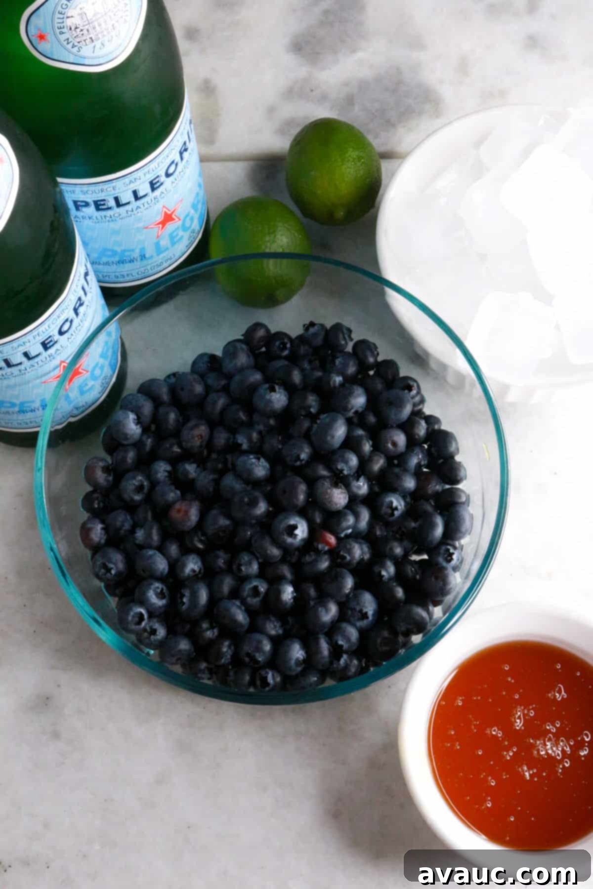 Vibrant Berry Refresher 3 Two bottles of Pellegrino sparkling water, a bowl of fresh blueberries, two limes, a bowl with ice, and a small bowl with honey, all ready for mocktail preparation.