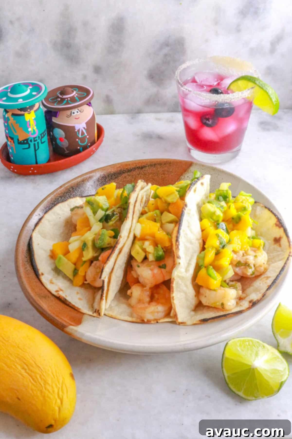 Zesty Shrimp Tacos with Tropical Mango Salsa 2 A plate with three shrimp tacos with mango salsa and a sparkling blueberry mocktail behind it