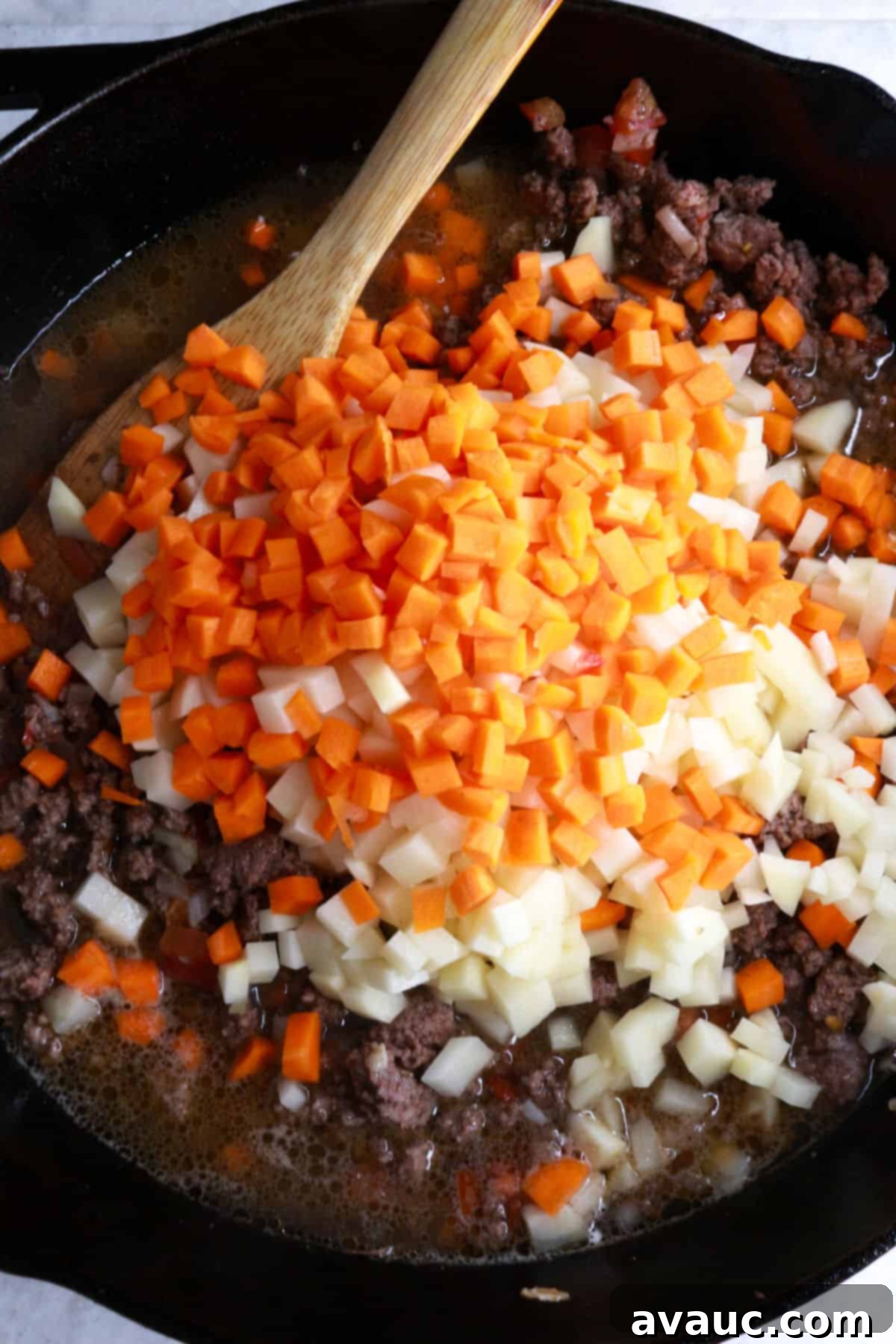 Diced carrots and potatoes being added to the ground beef mixture in a skillet.