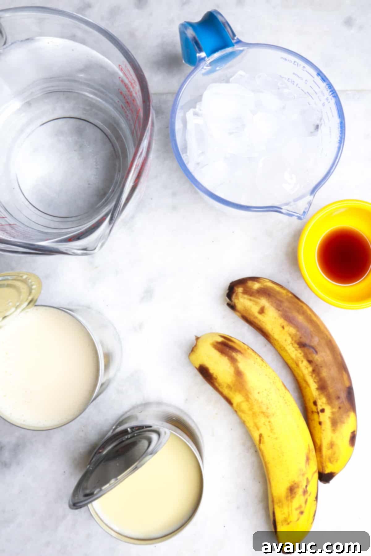 Fresh Banana Drink 3 A collection of ingredients for Agua de Plátano, including fresh ripe bananas, measuring cup of water, evaporated milk, condensed milk, vanilla extract, and ice