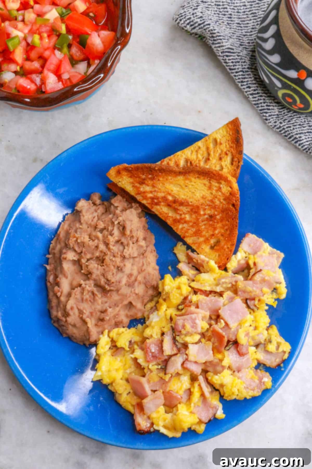 Classic Ham and Egg Scramble 2 Overhead view of a plate with huevos con jamon, refried beans and toast with a bowl of Pico de Gallo and a cup of coffee in the background