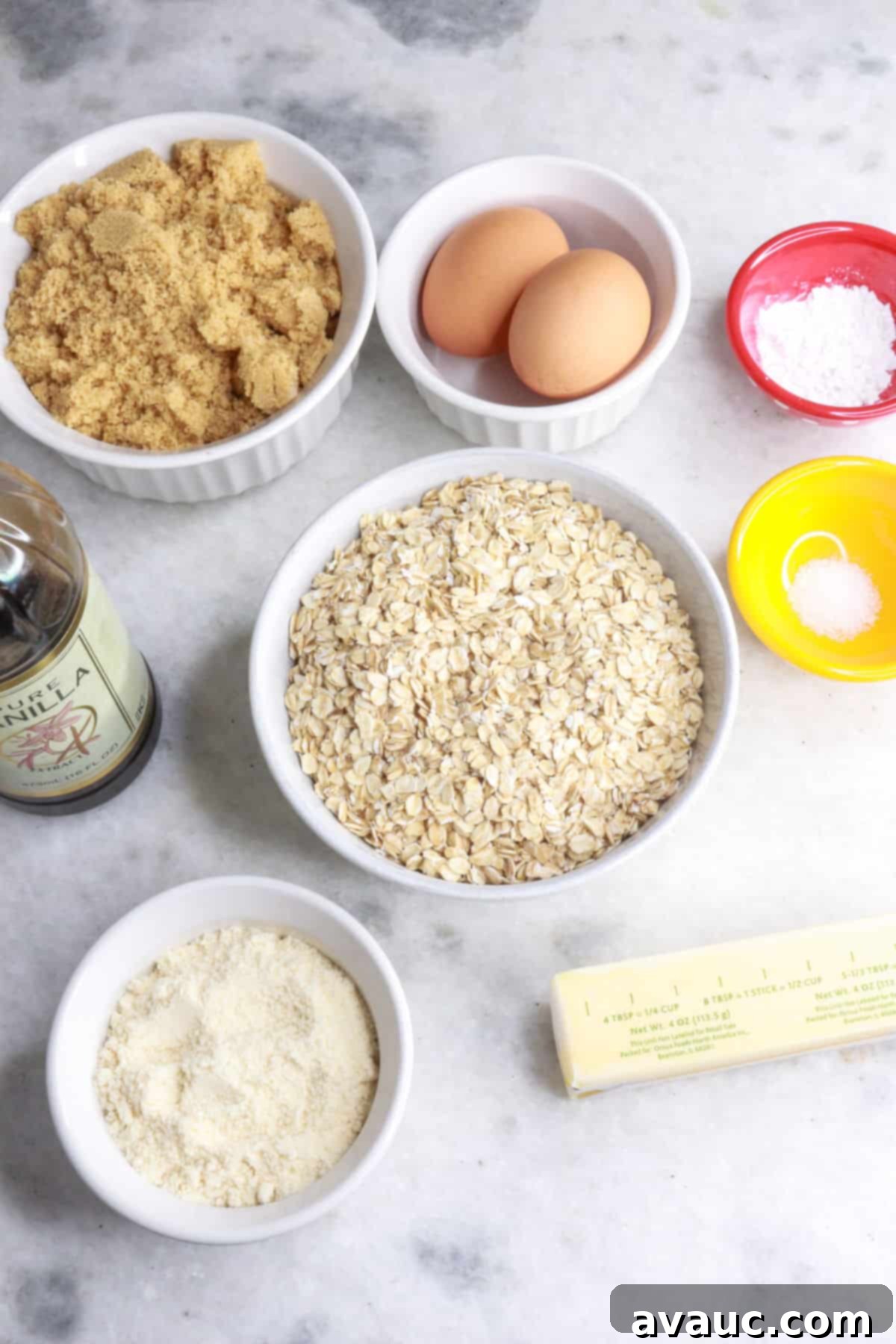 A collection of cookie ingredients laid out: a bowl of brown sugar, two eggs, small bowls with baking powder and salt, a bowl of old-fashioned oats, a stick of butter, coconut flour in a bowl, and a bottle of vanilla extract.