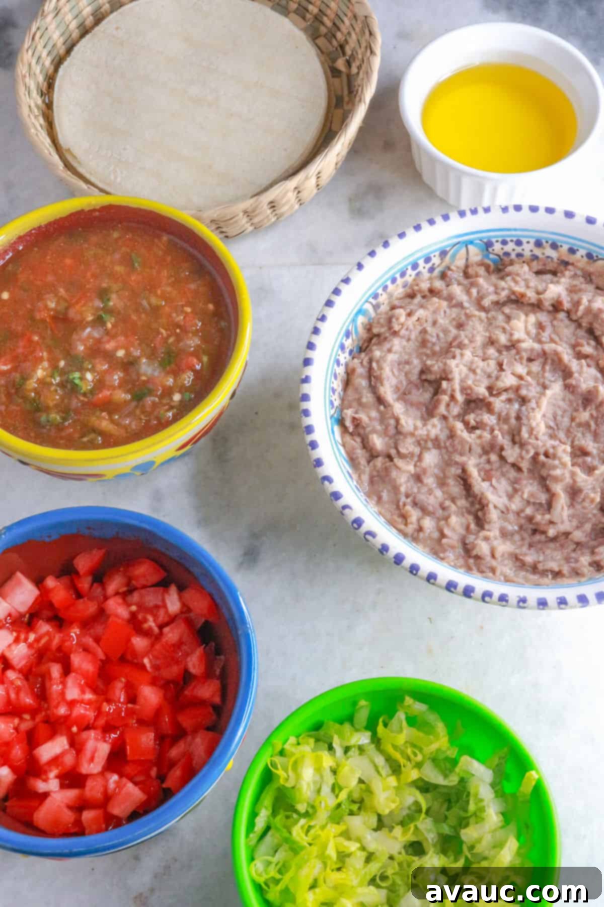 Golden Crispy Bean Tacos 3 A basket with corn tortillas, a bowl with oil, a bowl with jalapeño salsa, a bowl with refried beans, another with diced tomatoes and another with shredded lettuce