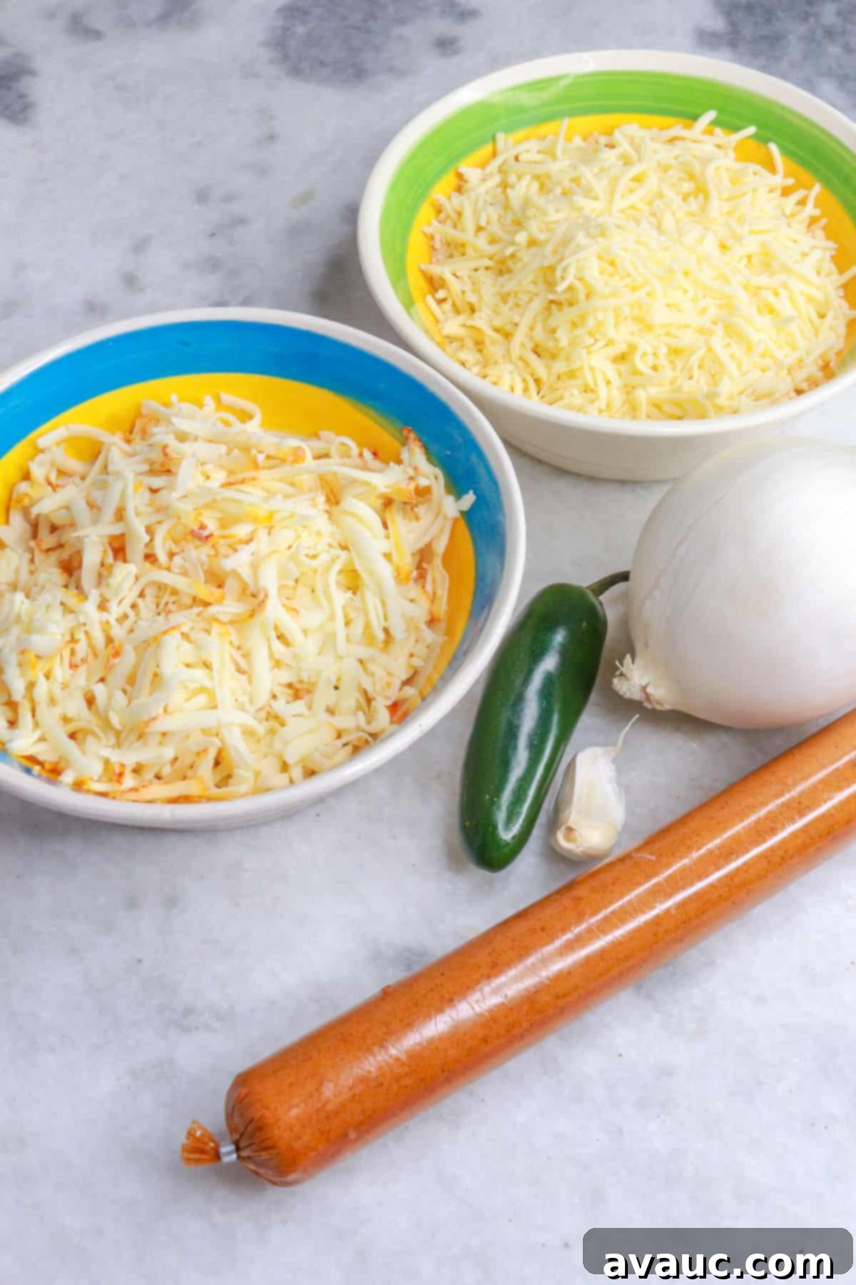 Spicy Chorizo Queso Dip 3 Two bowls of shredded Mexican cheese, one fresh jalapeño, a whole clove of garlic, a white onion, and uncooked Mexican chorizo, ready for choriqueso preparation.