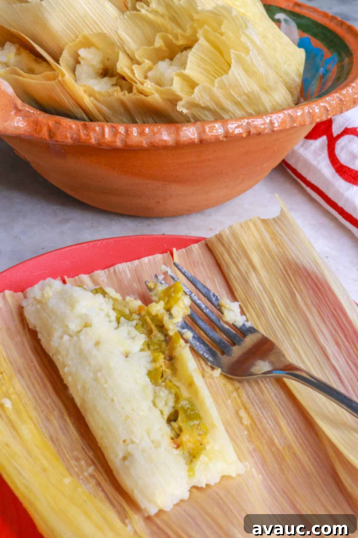 An open green chile and cheese tamal on a plate with a bowl of tamales behind it