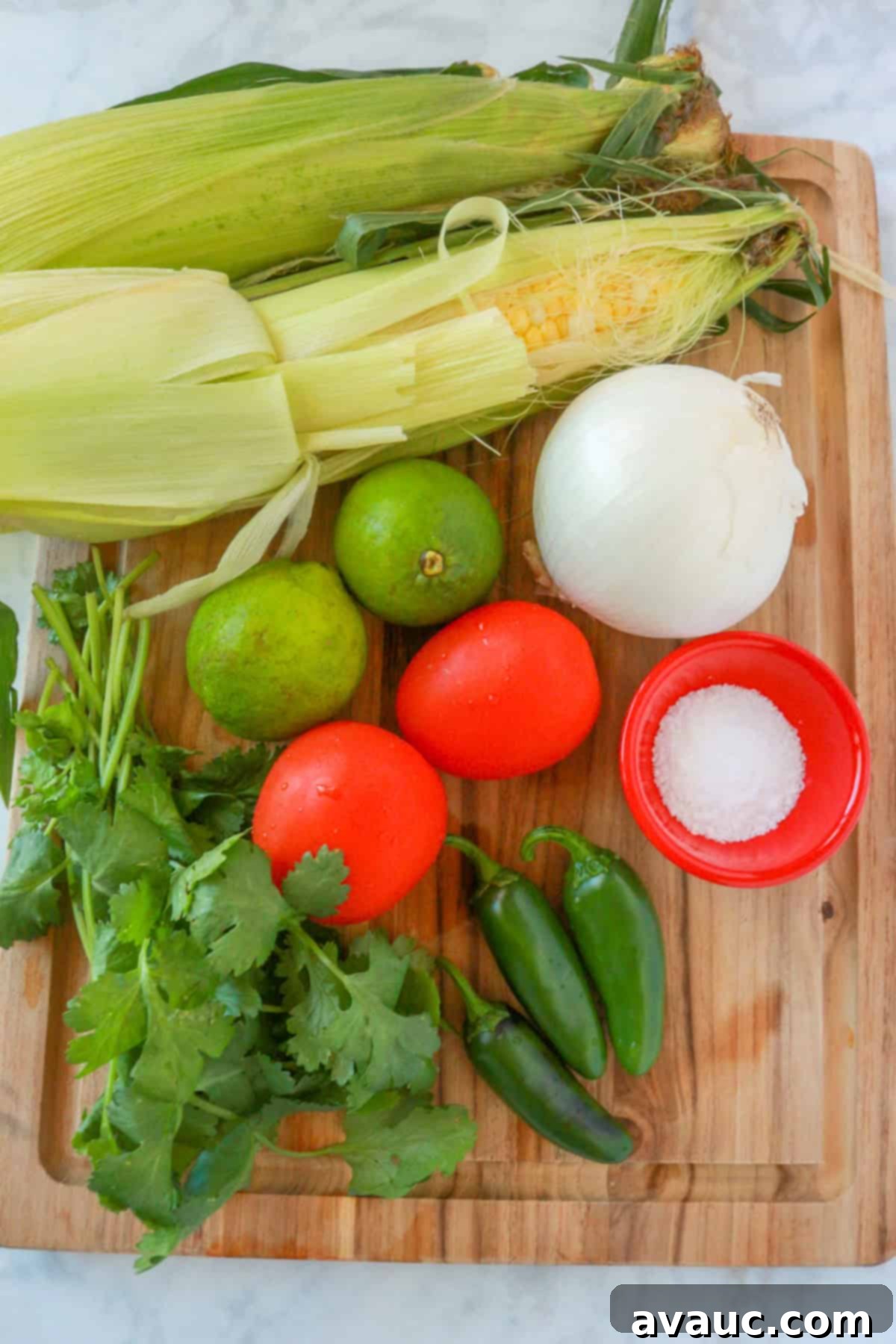 Fresh ingredients laid out on a wooden cutting board: ears of corn, bright green limes, a white onion, a small bowl of salt, ripe Roma tomatoes, vibrant jalapeños, and a bunch of fresh cilantro.