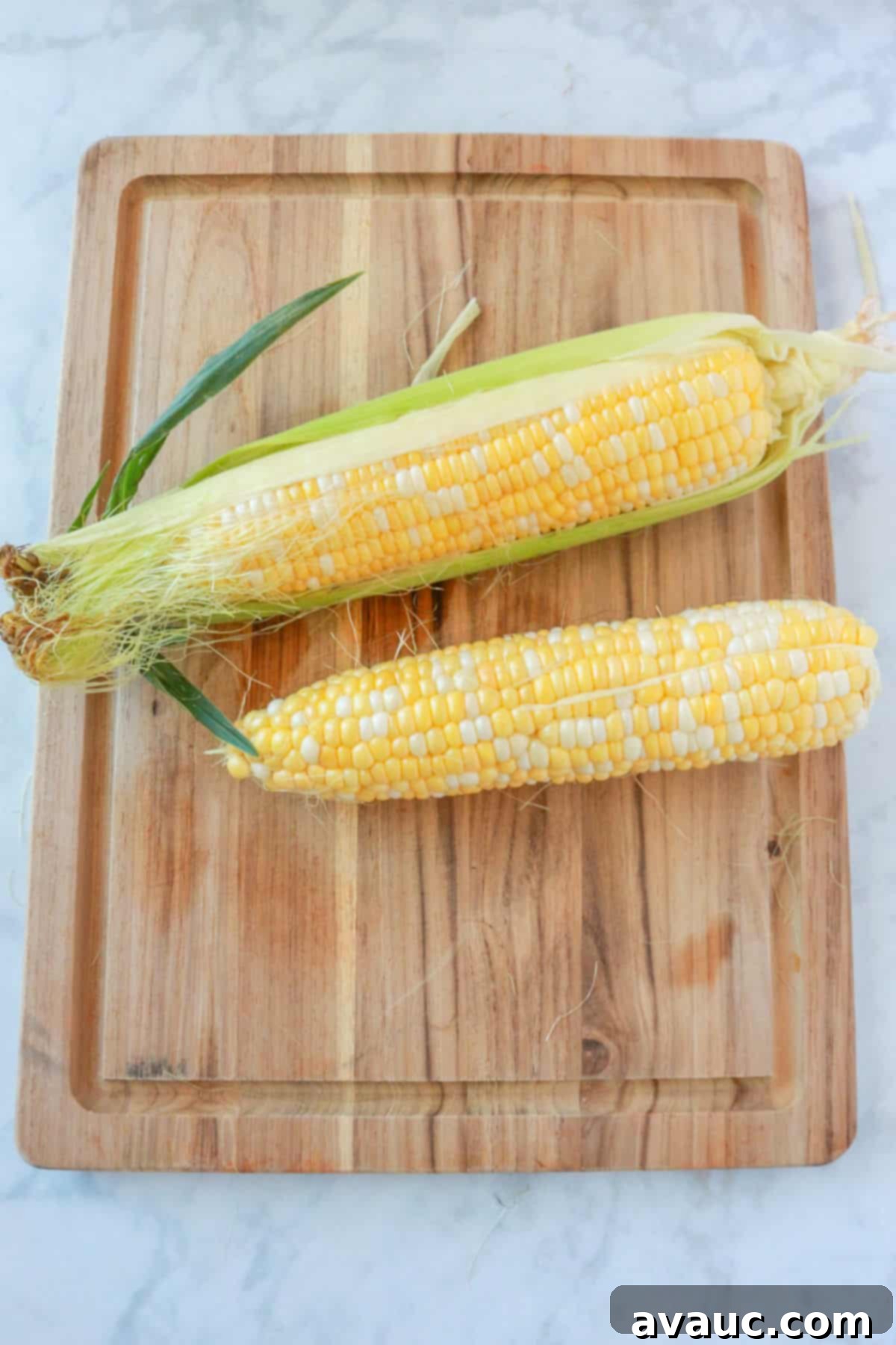 Two fresh ears of corn on a wooden cutting board, ready for preparation.