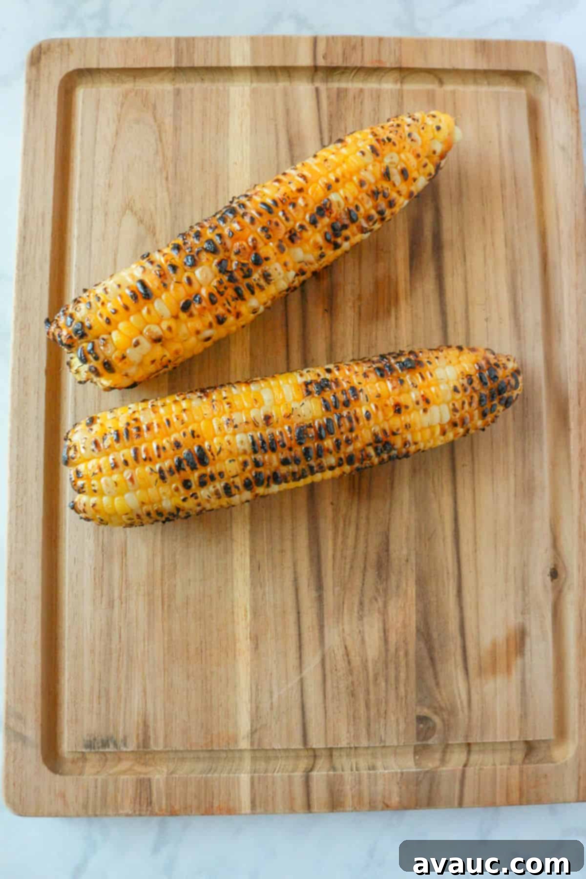 Two roasted ears of corn resting on a wooden cutting board, showing slight charring on the kernels.