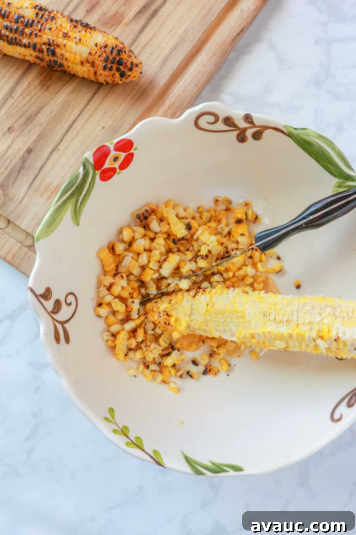 A sharp knife cutting roasted corn kernels off the cob into a large mixing bowl.