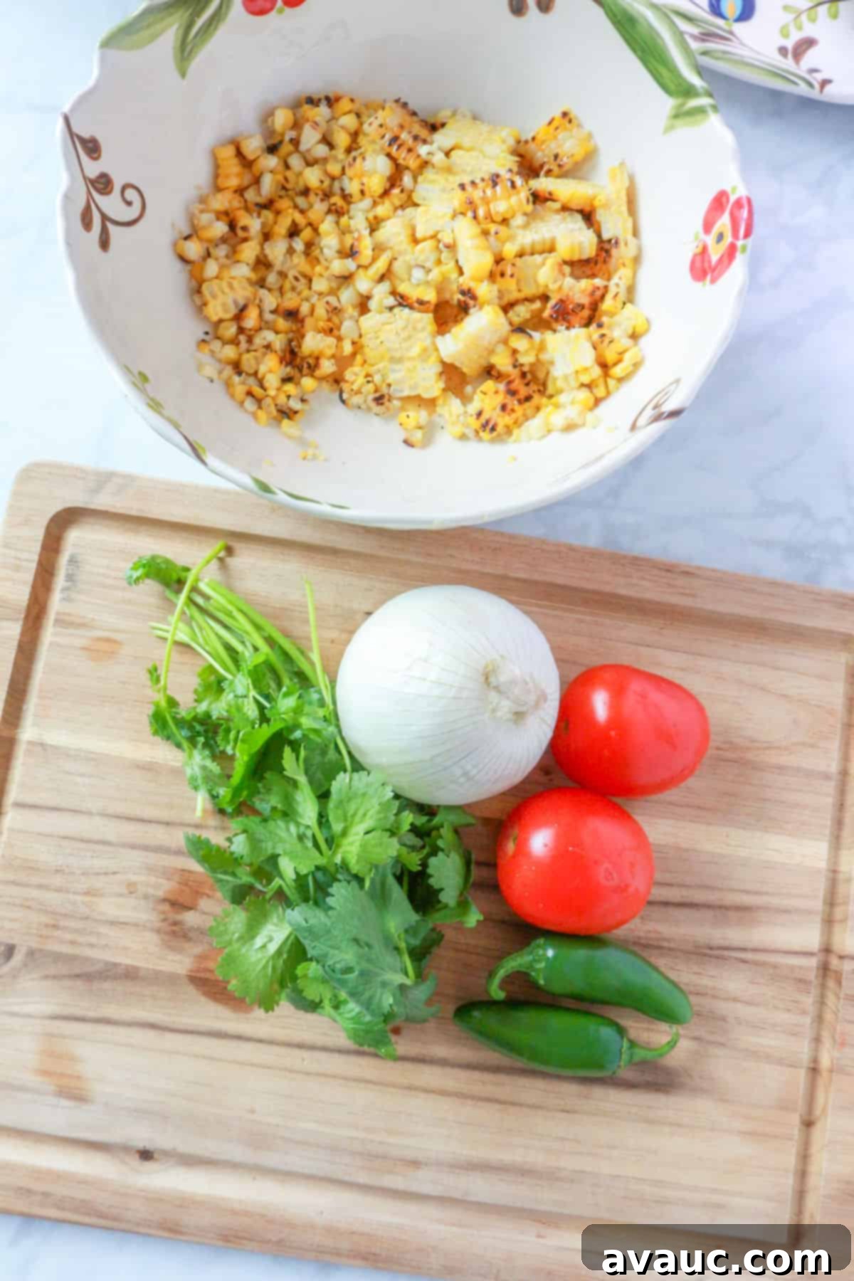 A wooden cutting board with fresh cilantro, a white onion, two Roma tomatoes, and two jalapeños, with a bowl of roasted corn kernels in the background.