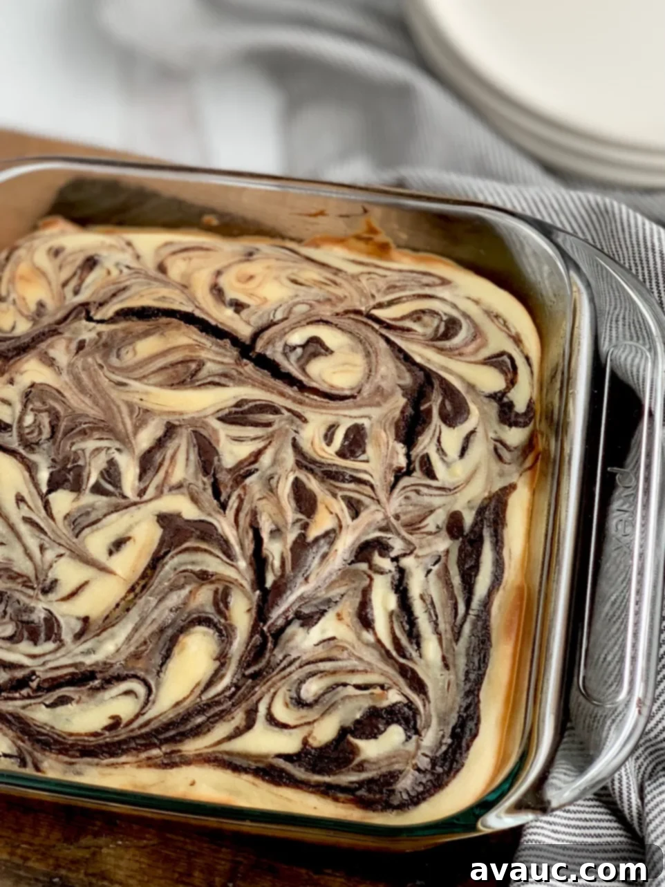 A glass 8x8-inch baking dish filled with a baked marbled brownie mixture with swirls of cream cheese and dessert plates in the background.