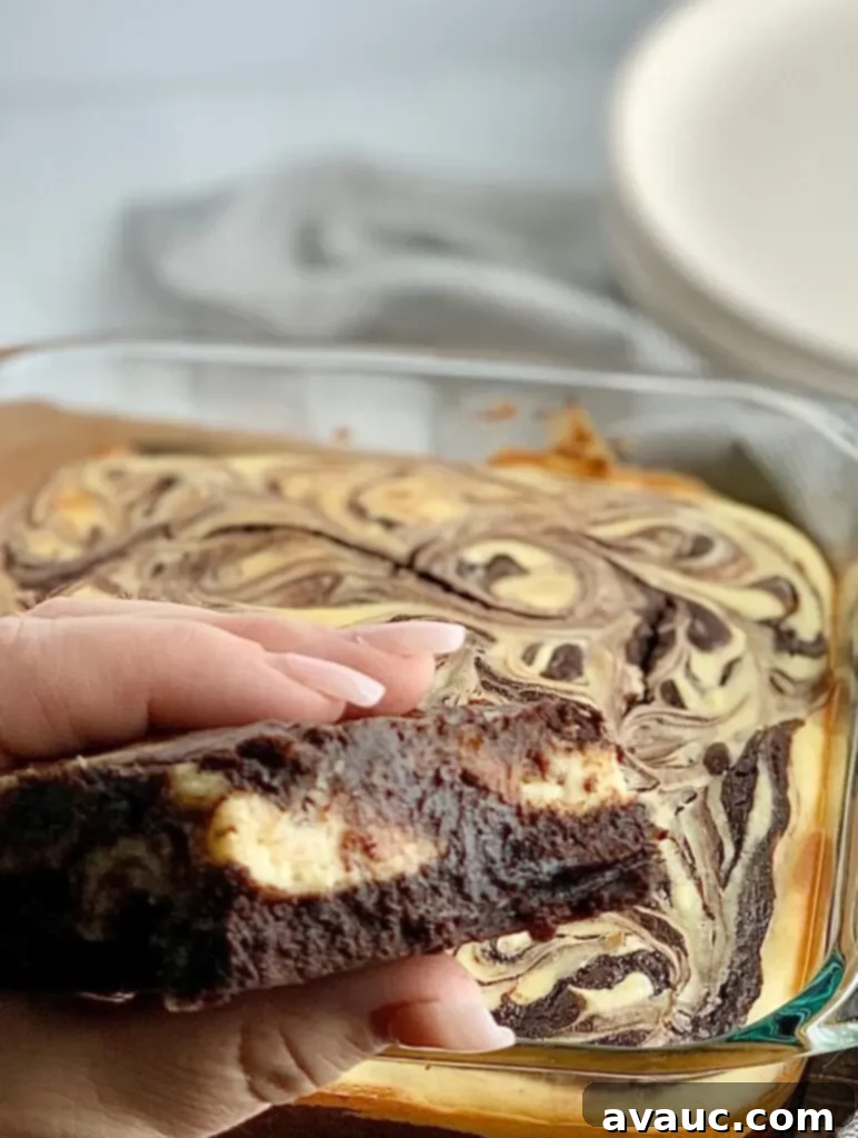 A person folding a cut out brownie with chunks of white cream cheese in it with an 8x8-inch baking dish filled with a baked marbled brownie mixture with swirls of cream cheese and dessert plates in the background.