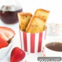 A serving of homemade French toast sticks in a striped red cup, garnished with fresh strawberries and a drizzle of syrup.