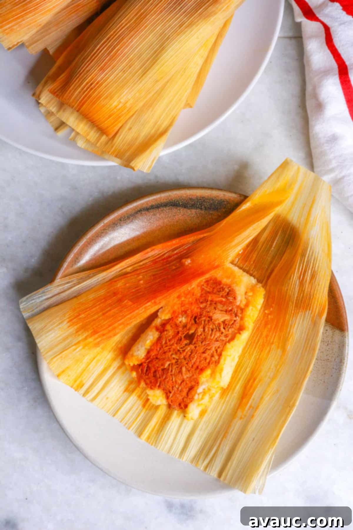 Red Chile Pork Tamales 2 Mexican tamales rojos on a plate and one tamal unwrapped on a brown plate