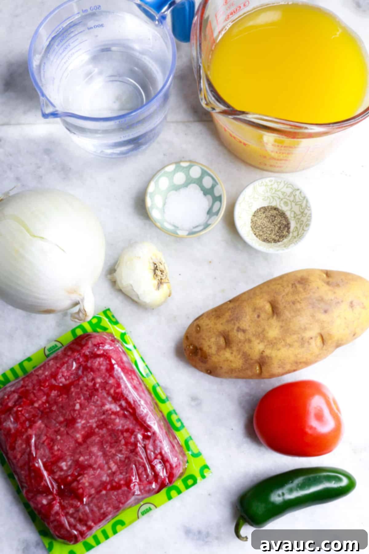 Jalapeño Ground Beef Stew 3 A flat lay of ingredients for Caldillo de Carne Molida: measuring cup with water, another with chicken broth, a whole onion, a small bowl with salt, another with pepper, a garlic bulb, a pound of ground beef, a potato, a tomato, and a jalapeño