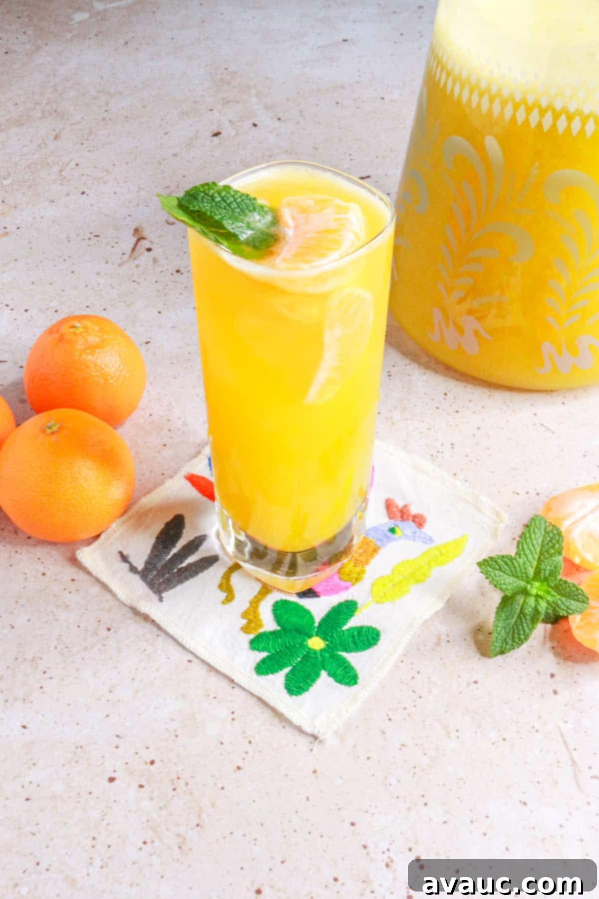 Refreshing Mandarin Mint Agua Fresca 10 Glass of mandarin Agua Fresca with mint on a coaster with a pitcher of the Agua Fresca behind it