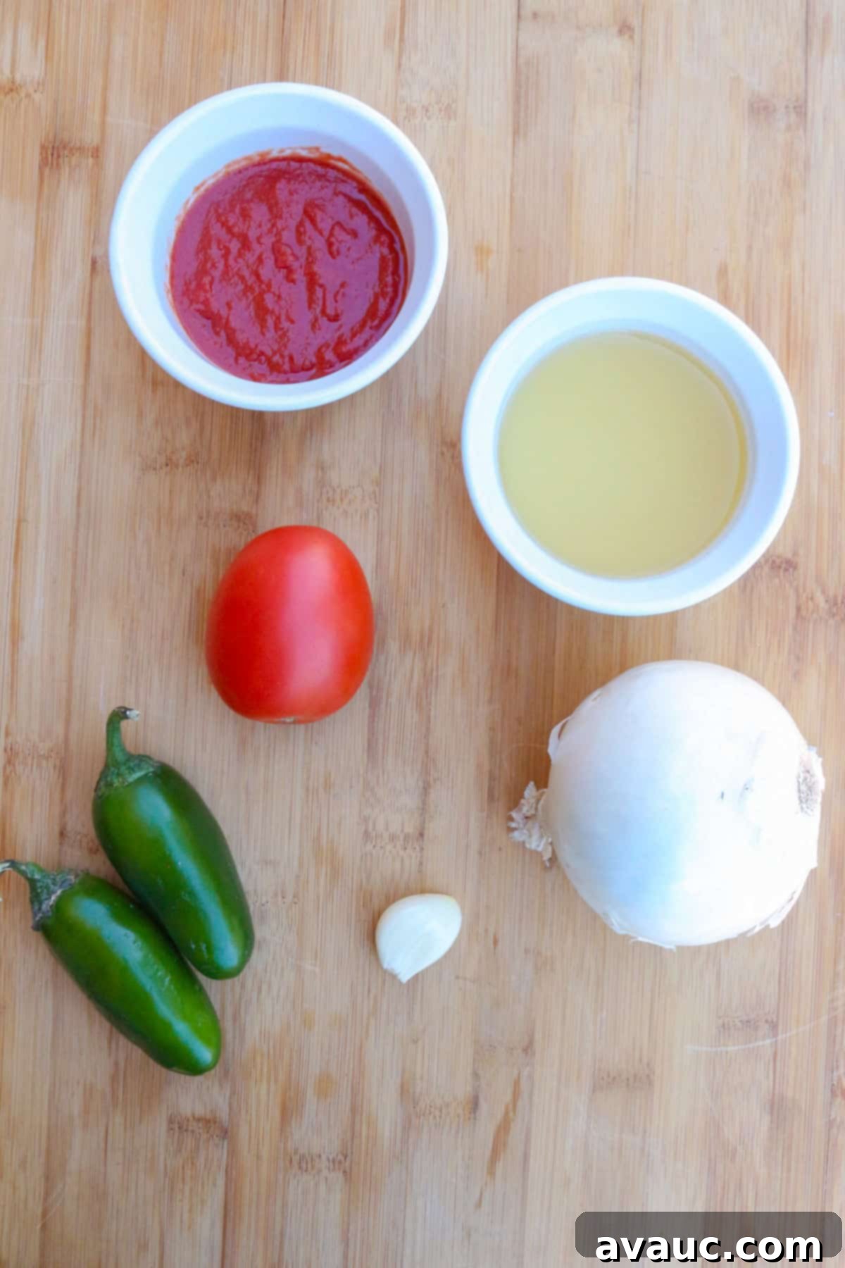 A small white bowl with tomato sauce, another with chicken broth, a Roma tomato, 2 jalapeños, one garlic clove and a white onion on a cutting board, ready for chunky salsa preparation