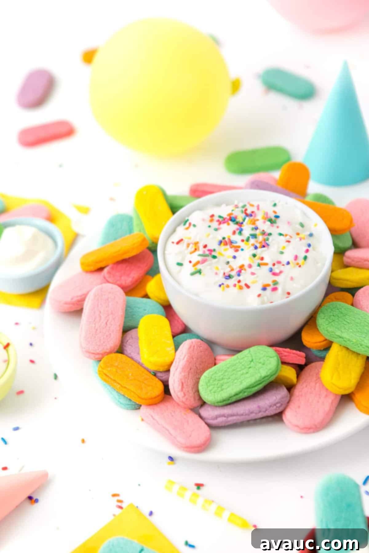 Plate of rainbow sprinkle sugar cookies with a bowl of cream cheese frosting in a party scene.