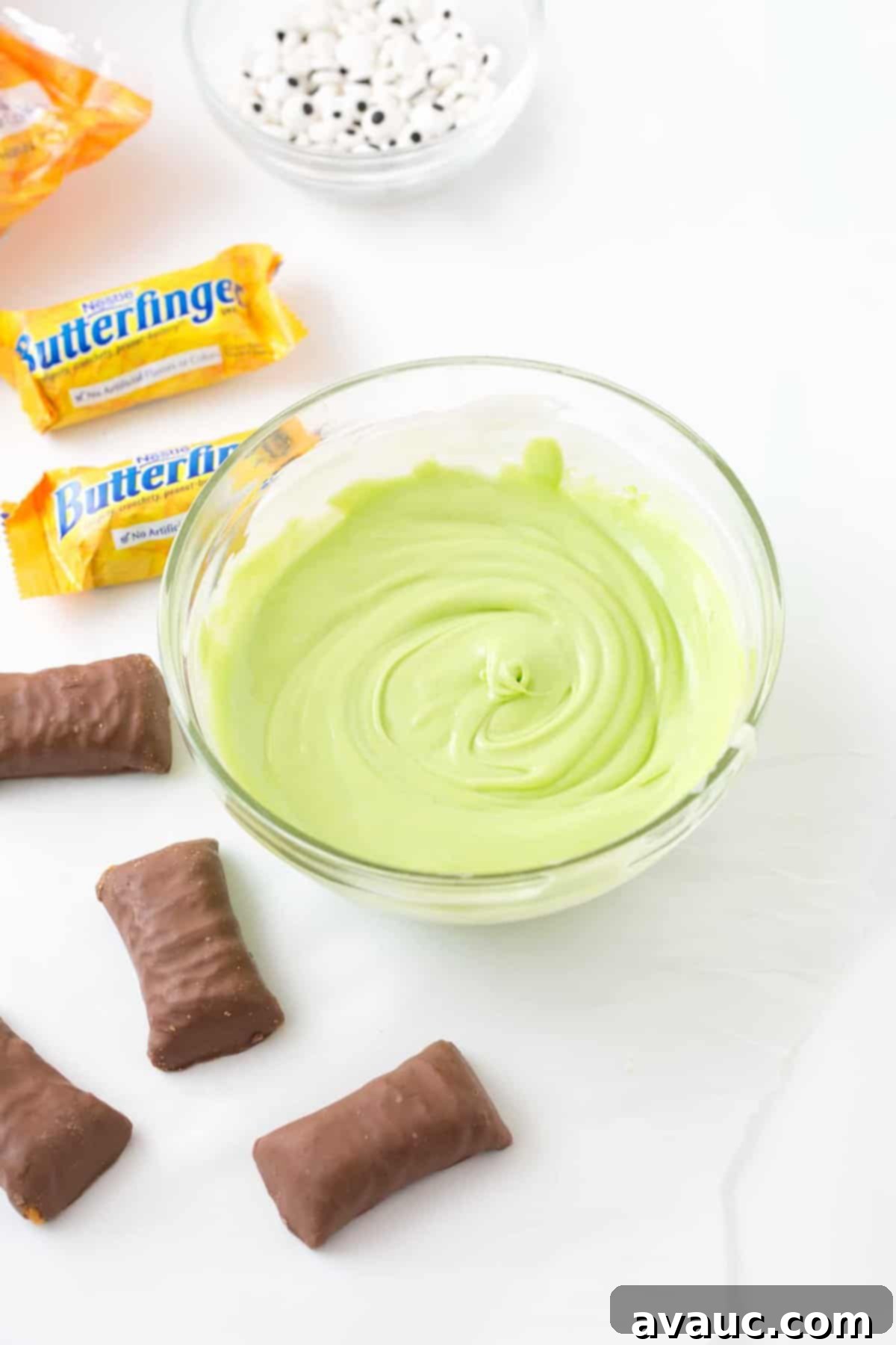Bowl of melted green almond bark with Butterfinger candy bars and a bowl of candy eyeballs off to the side.