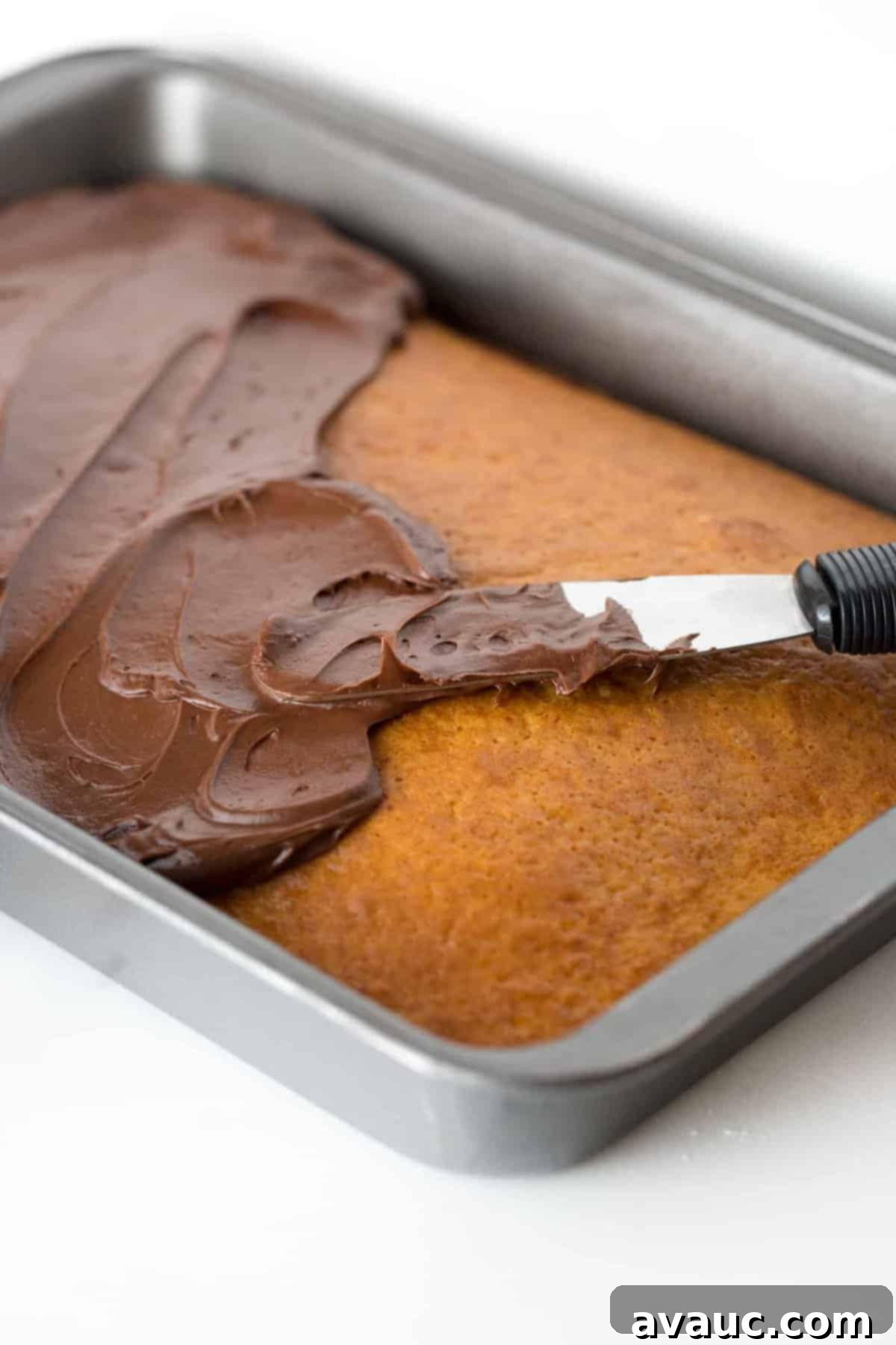 9x13 metal cake pan with yellow cake, spreading easy chocolate frosting on top with cake spreader.
