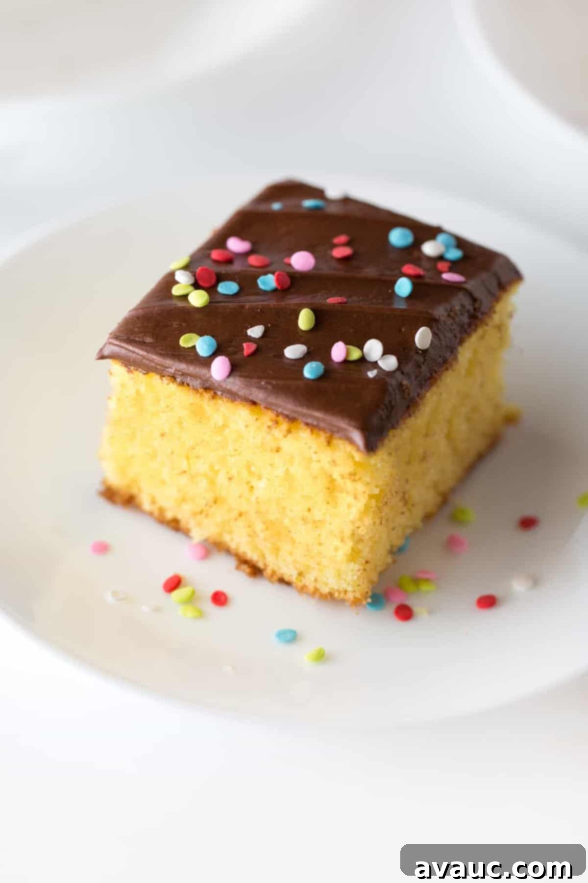 Square slice of yellow cake with easy chocolate frosting with rainbow sprinkle quins on top. On small white plate.