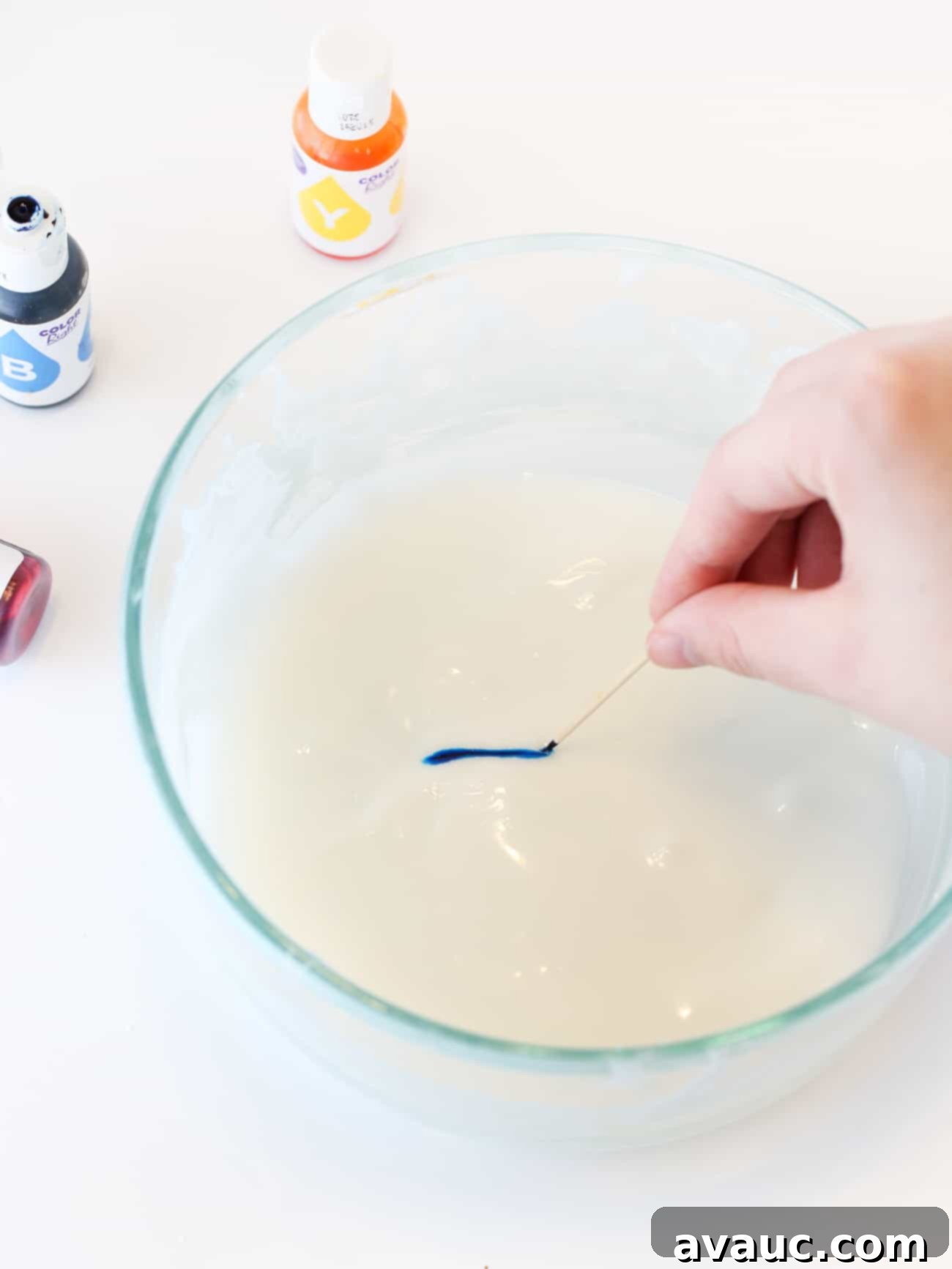 Hand holding toothpick that is adding blue food coloring to white cookie icing.