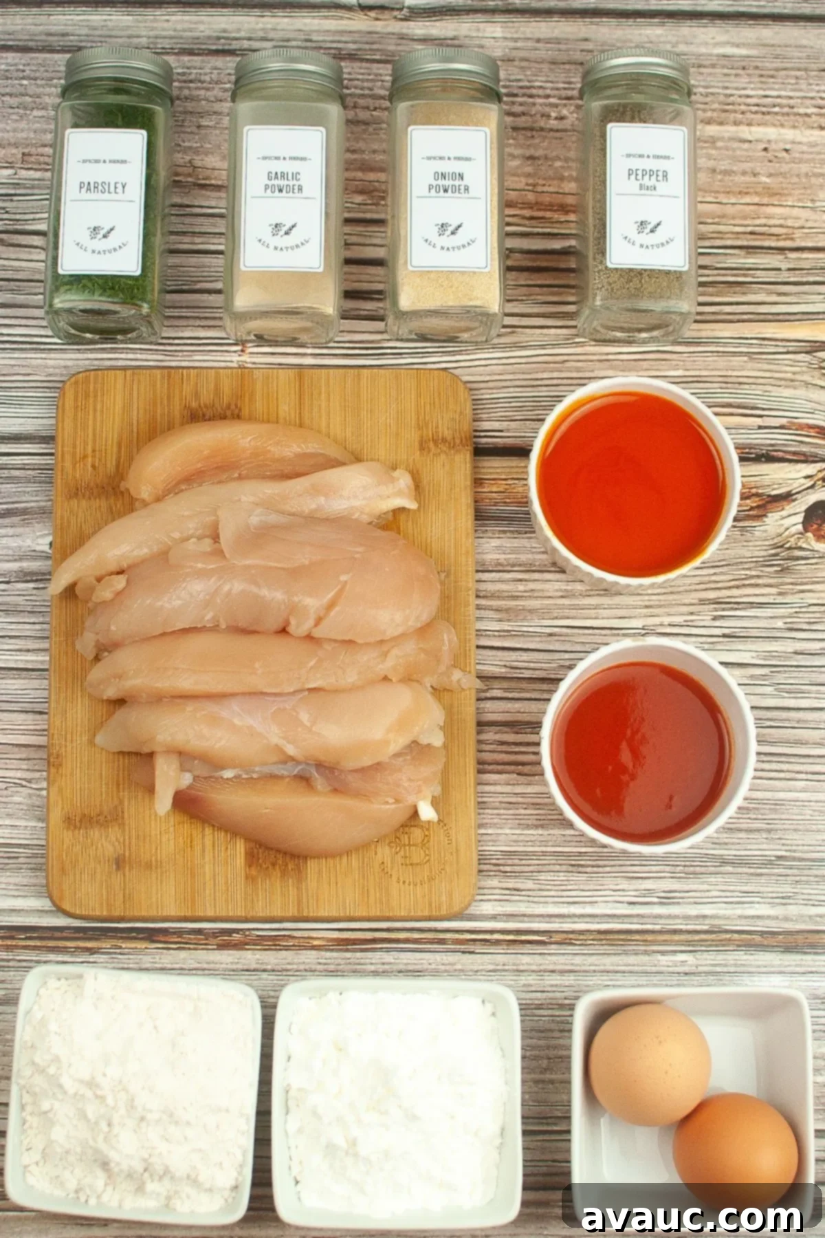 Ingredients laid out on a kitchen counter, including chicken tenders, gluten-free flour, cornstarch, various spices, eggs, hot sauce, and buffalo sauce.