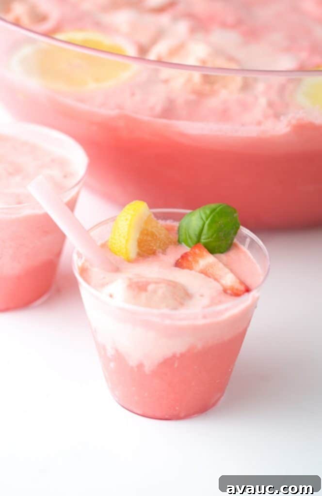 A single clear cup of Pink Strawberry Sherbet Punch, garnished with a lemon slice and basil, on a rustic wooden surface.