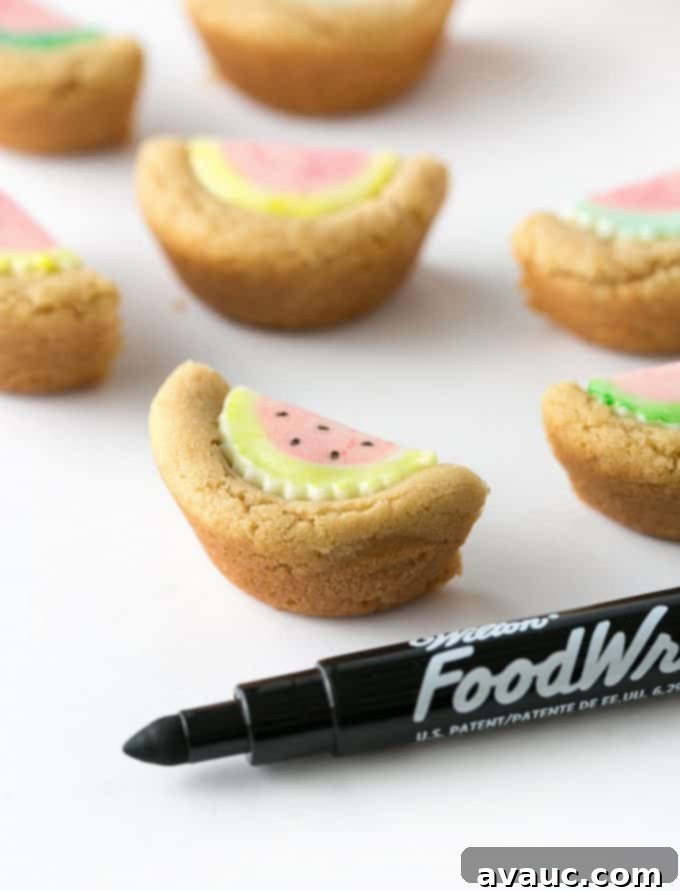 A close-up image showing the final step of decorating, where black food marker is used to draw seeds onto a watermelon cookie cup.