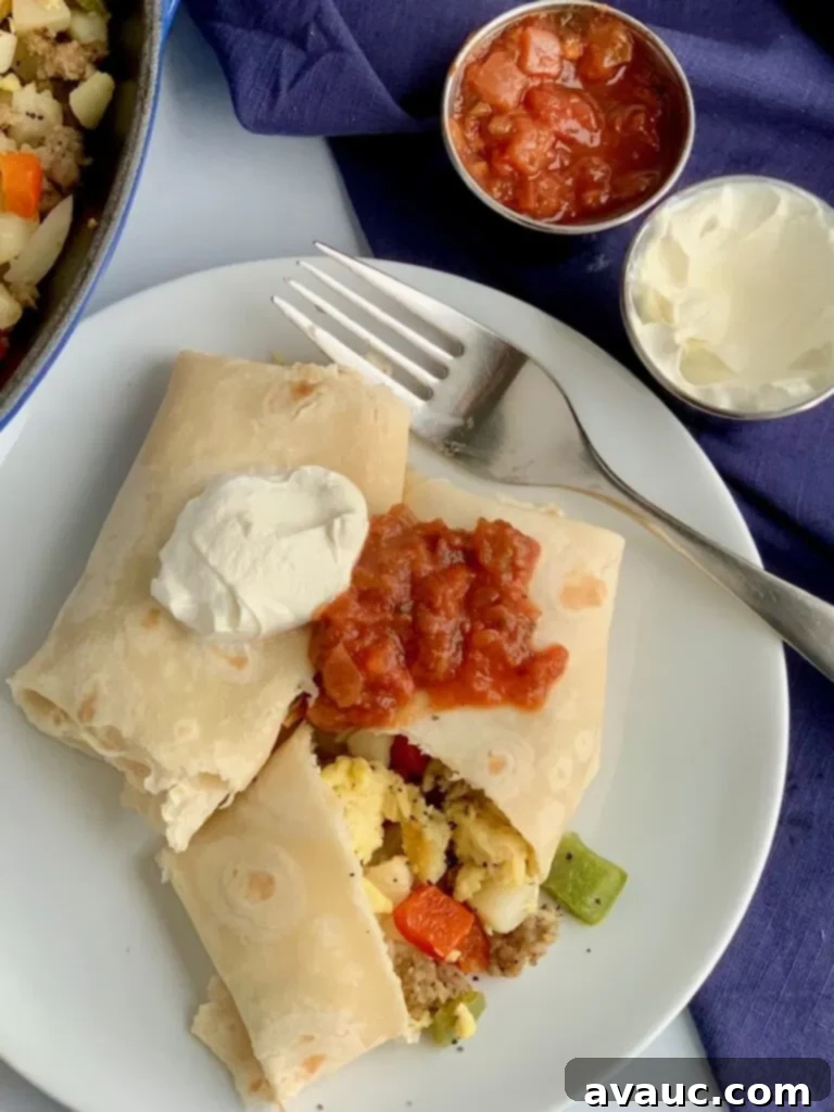 A white plate with 2 large breakfast burritos on it. One is cut open and you can see eggs, bell peppers, sausage, and hash browns. They are topped with dairy free sour cream and a dollop of salsa