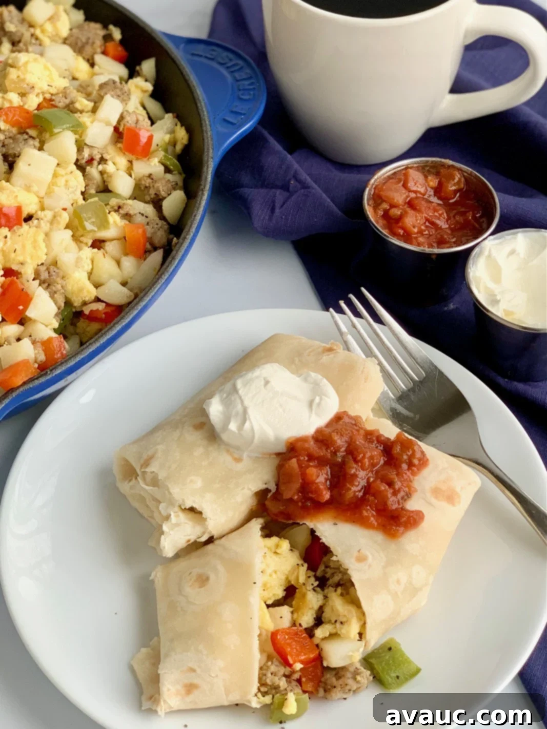 A white plate with 2 large breakfast burritos on it. One is cut open and you can see eggs, bell peppers, sausage, and hash browns. They are topped with dairy free sour cream and a dollop of salsa.