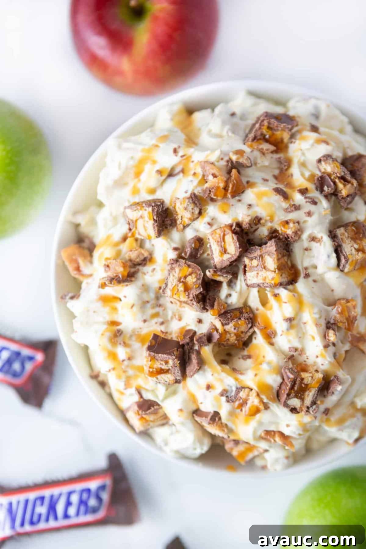 White bowl of apple snickers salad. A fluffy vanilla pudding dessert topped with chopped snickers candy bars and caramel sauce.