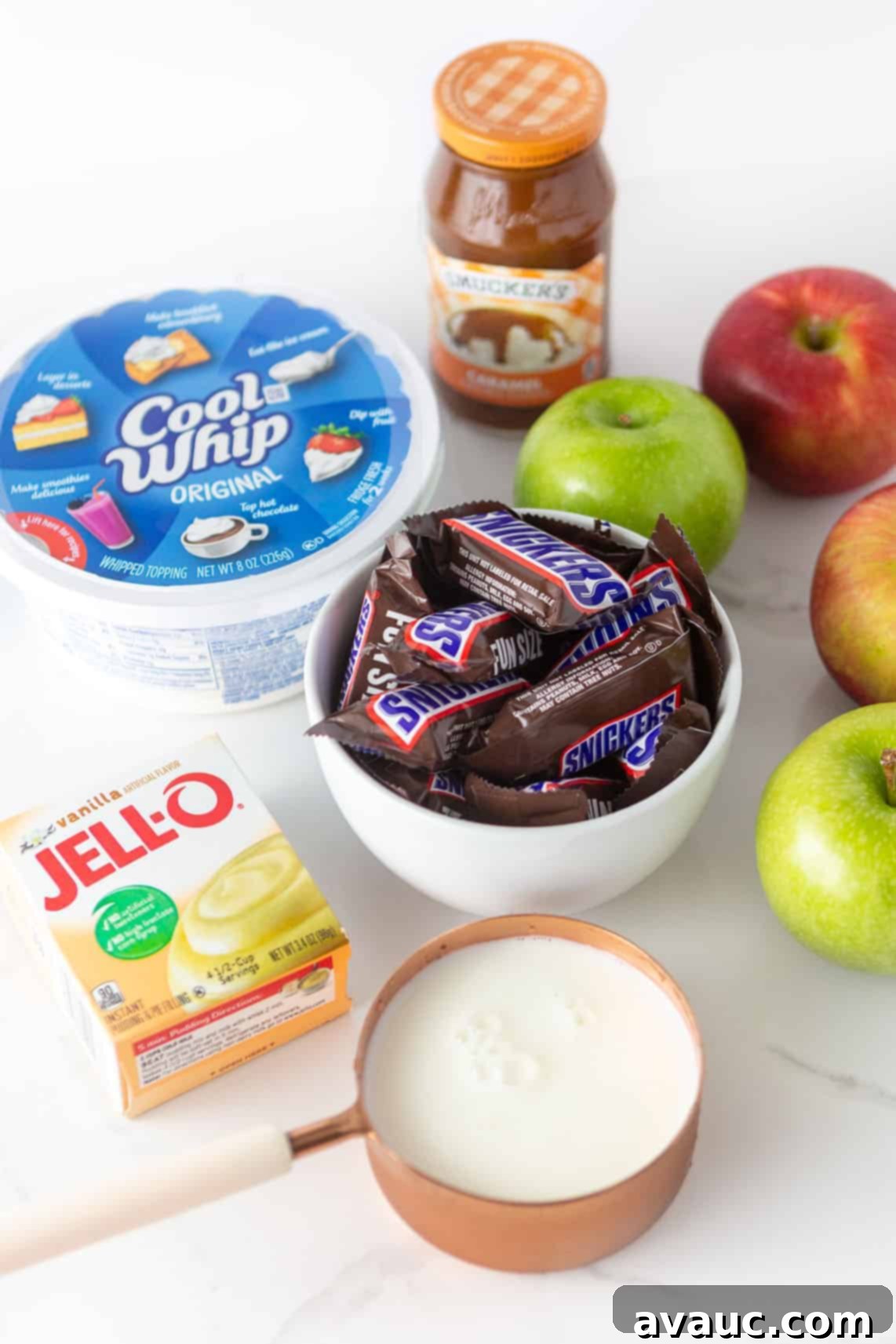 Ingredients for apple snickers salad; includes bowl of snickers, cool whip, a box of instant vanilla pudding mix, milk, apples, and a jar of Smuckers caramel sauce