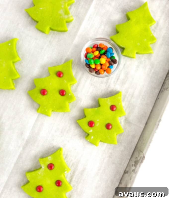 Effortless Christmas Tree Cookies 4 Unbaked green Christmas tree sugar cookies with red M&M ornaments pressed into their tops, arranged neatly on a baking pan, ready for the oven.