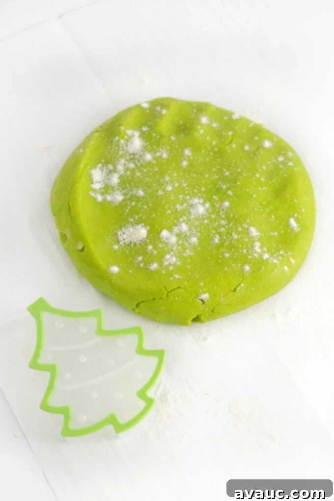 Effortless Christmas Tree Cookies 5 A ball of bright green sugar cookie dough resting beside a metal Christmas tree cookie cutter on a floured surface, ready for cutting.