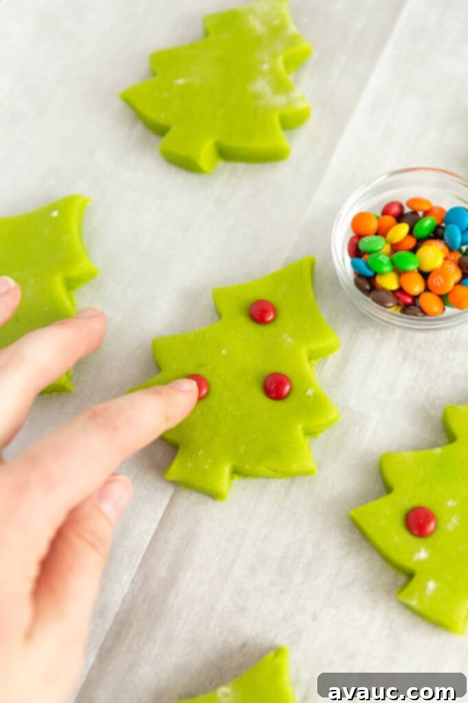 Effortless Christmas Tree Cookies 8 A person's finger gently pressing a mini red M&M candy into the soft green Christmas tree shaped dough on a baking sheet, preparing it for baking.