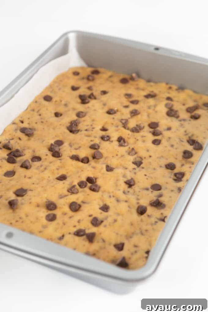 Irresistible Chocolate Chip Squares 8 Chocolate chip cookie dough being pressed into a 9x13 inch baking pan, lined with parchment paper.