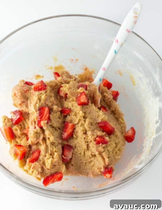 Cheesecake Filled Strawberry Cookies 3 Glass bowl filled with cookie dough, a separate bowl with sliced fresh strawberries, and a colorful confetti spatula on a white countertop, illustrating the preparation process.