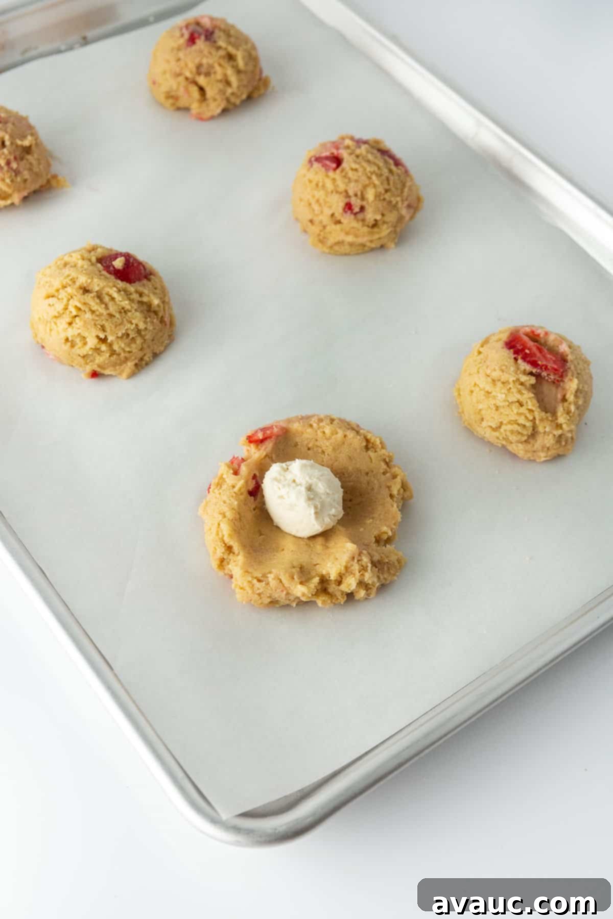 Cheesecake Filled Strawberry Cookies 6 A hand gently wrapping strawberry cookie dough around a creamy cheesecake filling ball on a baking sheet, preparing the cookies for baking.