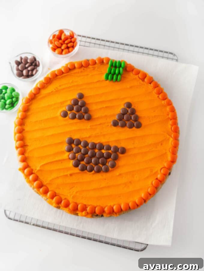 Spooktacular Easy Pumpkin Cookie Cake 8 round chocolate chip cookie cake with orange frosting and m&m's pressed on top to create a smiling Halloween jack-o-lantern pumpkin face