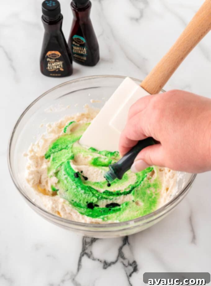 Festive Christmas Tree Sugar Cookie Bars 9 A hand adding green food coloring drops to white frosting in a bowl, mixing it to create a vibrant green hue for the Christmas tree sugar cookie bars.