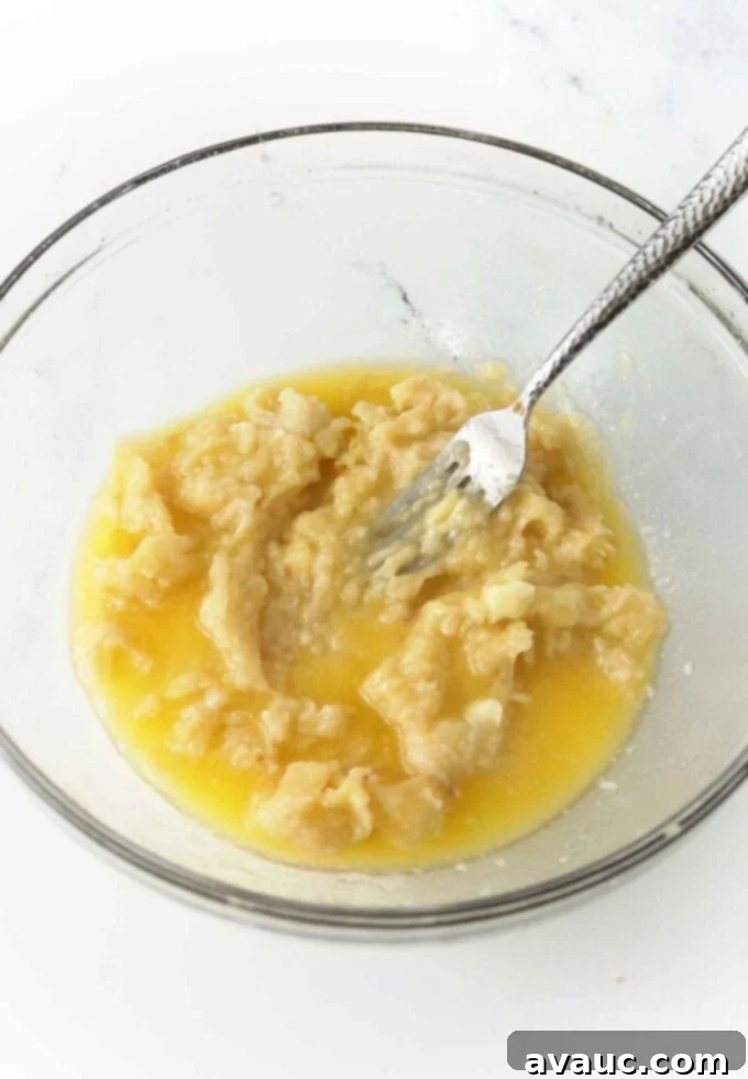Glass bowl containing mashed bananas and melted butter, ready for mixing to create a smooth batter.