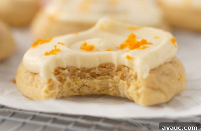 Glazed Orange Sugar Cookies 3 Two orange sugar cookies, one with frosting, placed on a white surface, with another cookie blurred in the background.