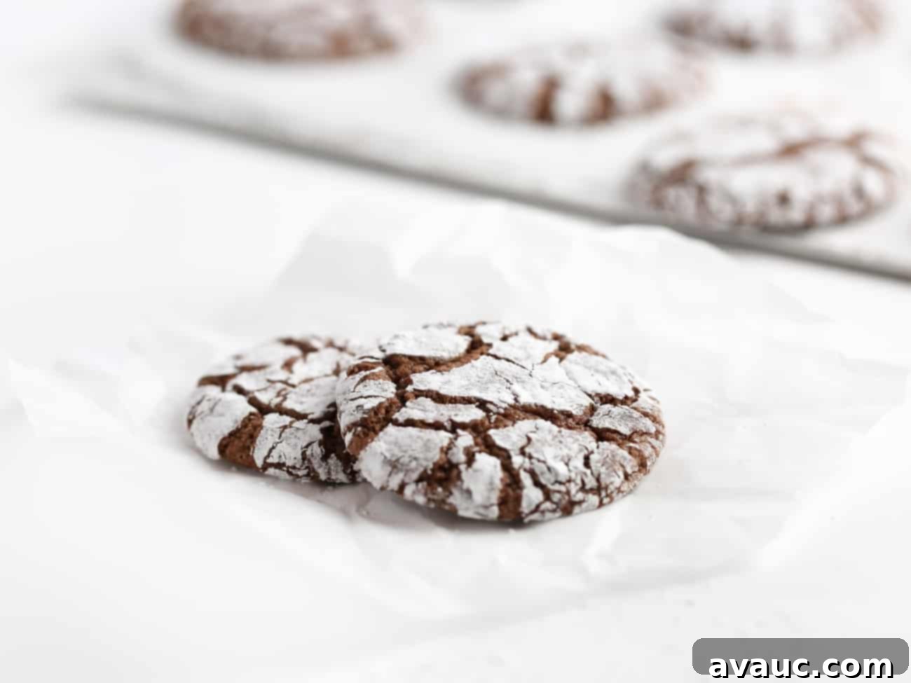 Two crackle-top chocolate brownie cookies in the foreground, with a baking tray of more cookies blurred in the background, emphasizing their homemade appeal.