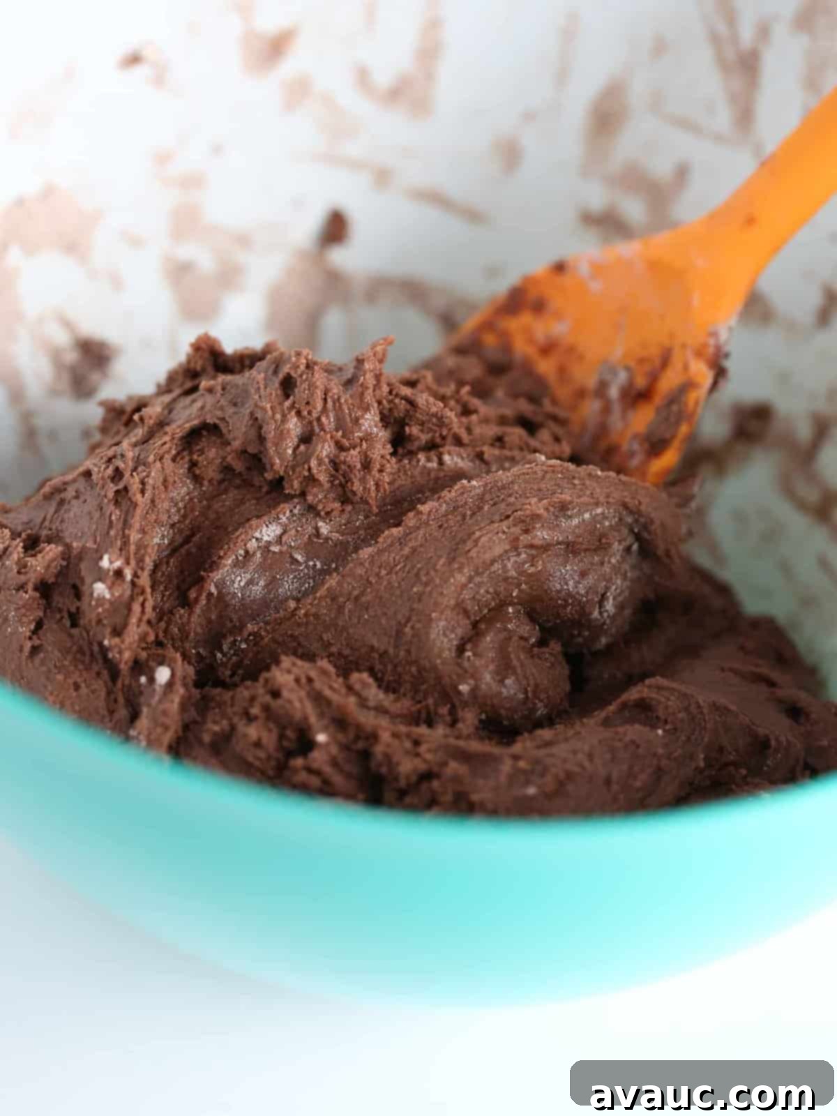 Orange silicone spatula stirring thick chocolate cookie dough for chocolate brownie cookies in a bowl, showcasing the density of the mixture.