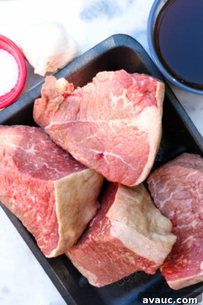 Raw beef chuck roast cut into large, even pieces, surrounded by beef broth, whole peeled garlic cloves, half an onion, and kosher salt on a rustic cutting board, prepared for cooking.
