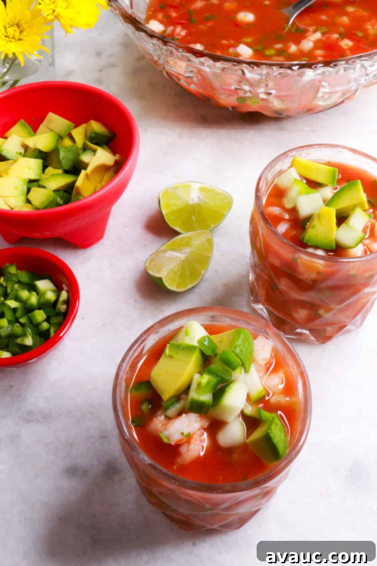 Vibrant Mexican Shrimp Cocktail 10 Two glasses filled with Coctel de Camarones, with a larger glass bowl of the cocktail behind them, surrounded by smaller bowls of diced avocado and diced jalapeño.
